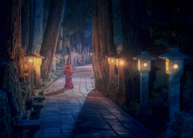 Paisaje nocturno y místico del cementerio budista de Okunoin en Japón, mostrando un camino de piedra entre árboles milenarios, luces cálidas de faroles tradicionales y la figura solitaria de una persona con kimono rojo.