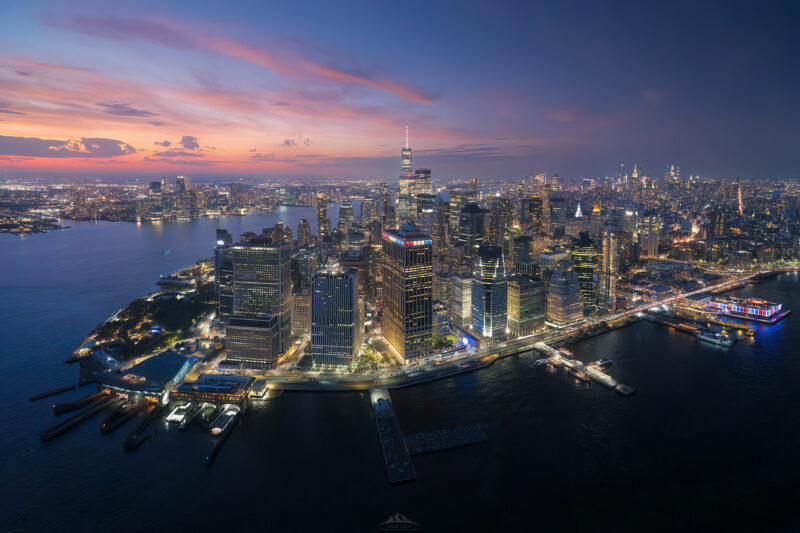 Fotografía aérea de Lower Manhattan en Nueva York durante el crepúsculo. Vista panorámica nocturna del distrito financiero iluminado, Battery Park y el río Hudson bajo un cielo de tonos rosados y azules.