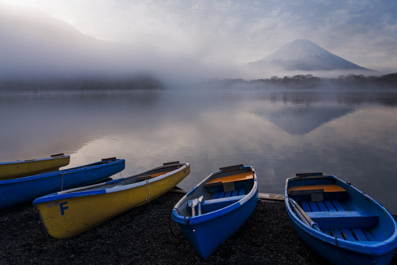 Fotografía de paisaje al amanecer. En primer plano, varias barcas de madera de colores azul y amarillo amarradas en la orilla de un lago. Al fondo, la imponente silueta del Monte Fuji emerge entre una densa capa de niebla blanca, reflejándose perfectamente en las aguas tranquilas y cristalinas.