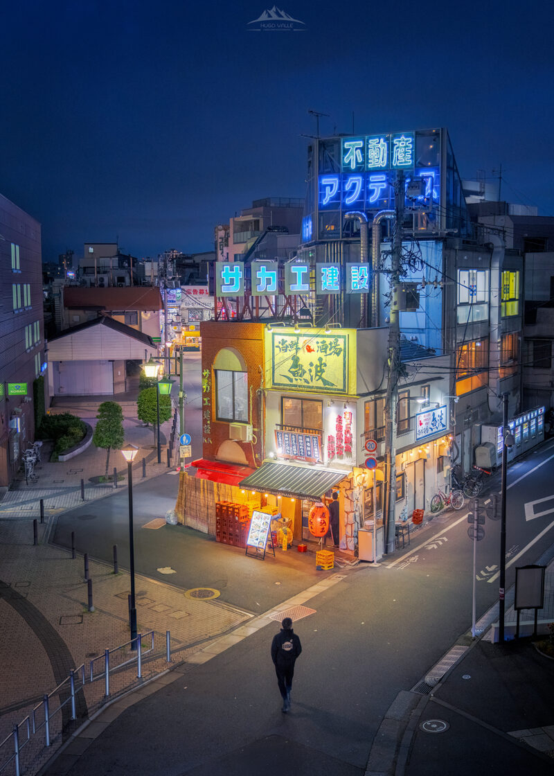 Vista aérea nocturna de una calle en Japón donde una persona camina sola hacia un edificio esquinero iluminado con letreros de neón brillantes en azul y amarillo, bajo un cielo crepuscular oscuro.