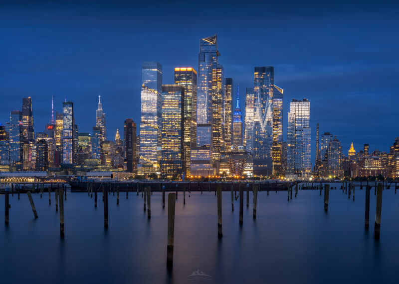 Postes de muelle de madera en primer plano con el horizonte iluminado de Midtown Manhattan y Hudson Yards al fondo, incluyendo el Empire State Building iluminado de azul y The Vessel, bajo un cielo de hora azul.