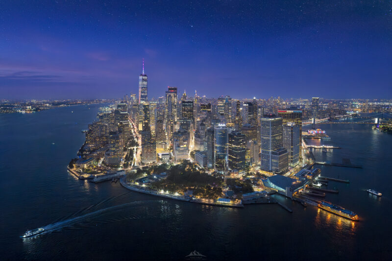 Fotografía aérea de alta resolución del Bajo Manhattan, Ciudad de Nueva York, durante el crepúsculo (hora azul). El horizonte del Distrito Financiero está totalmente iluminado contra un cielo nocturno azul profundo lleno de estrellas visibles. La prominente aguja del One World Trade Center se eleva sobre otros rascacielos. Battery Park está en primer plano, limitando con el agua oscura del río Hudson y el East River, donde los barcos dejan estelas de luz. Paisaje urbano nocturno icónico de NYC.