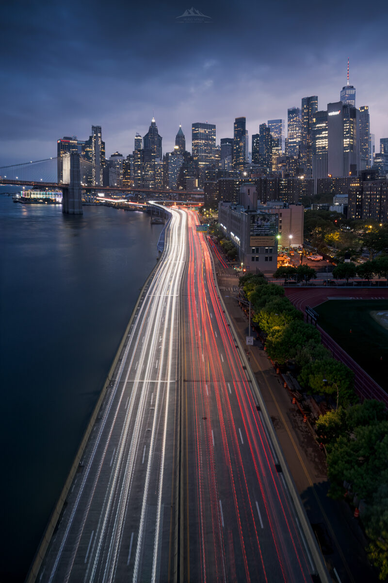 Fotografía nocturna de Nueva York con exposición prolongada en la autopista FDR Drive, mostrando estelas de luces de coches junto al East River, el Puente de Brooklyn y los rascacielos iluminados de Lower Manhattan.