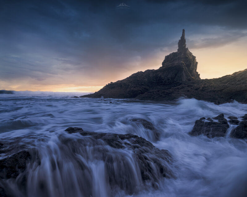 Fotografía de larga exposición del amanecer en el Peñón de Frexulfe, playa de Frexulfe (Navia, Asturias). Muestra el oleaje sedoso fluyendo sobre las rocas y un cielo enrarecido con tonos dorados y nubes de tormenta.