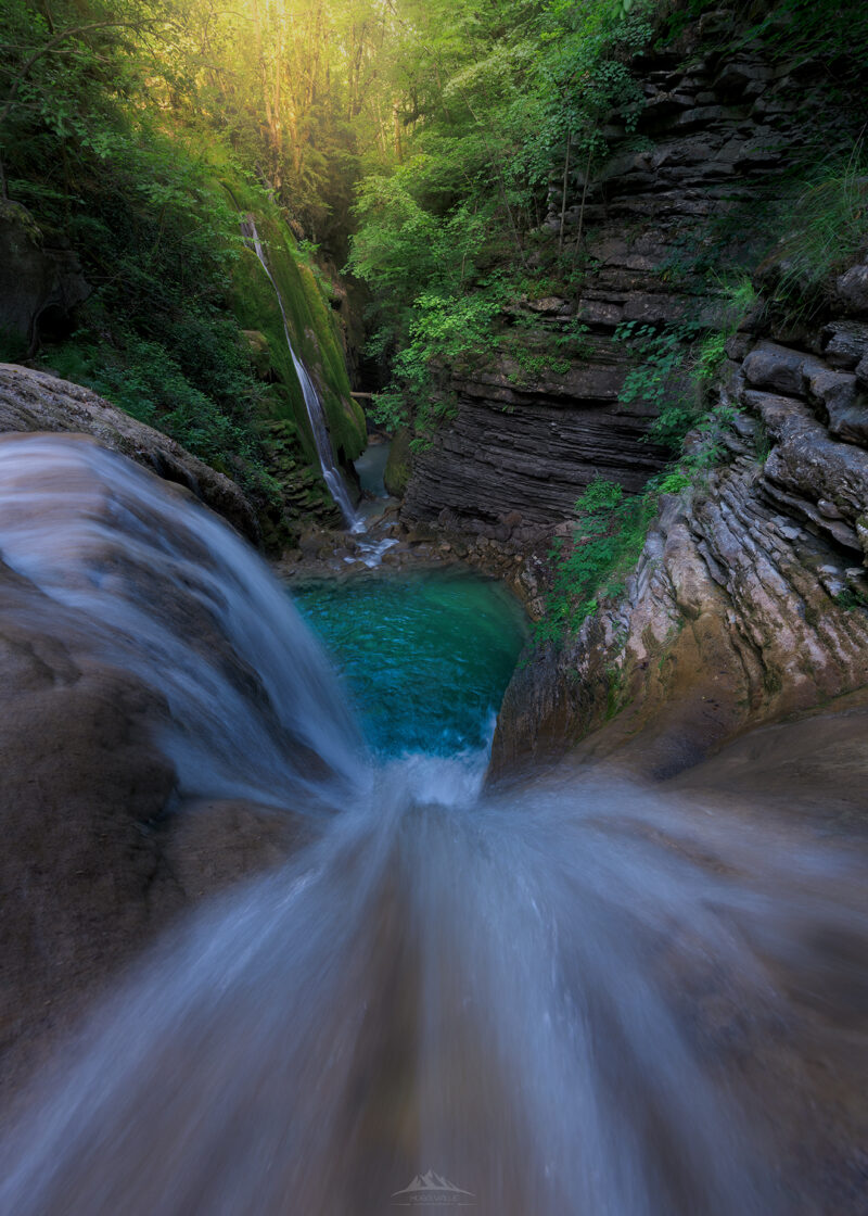 Vista en perspectiva superior del inicio del barranco de Forcos, donde una corriente de agua con efecto seda cae hacia una poza turquesa enmarcada por paredes de roca estratificada, vegetación verde y luz dorada al fondo.