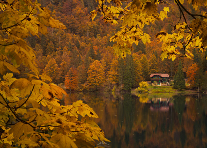 Cabaña de madera a orillas del Lago Fusine en los Alpes italianos durante el otoño, con el bosque de colores intensos reflejado en el agua.