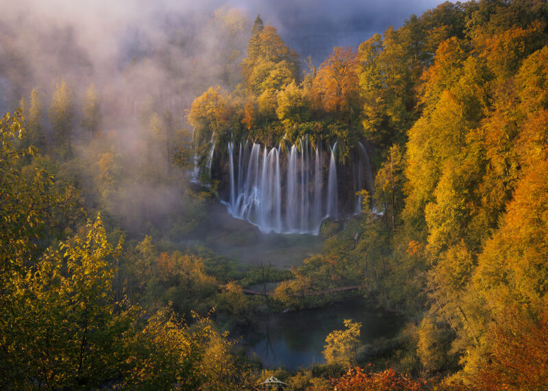 Cascada en los Lagos de Plitvice rodeada de un denso bosque otoñal con follaje dorado y bancos de niebla sobre el agua turquesa.